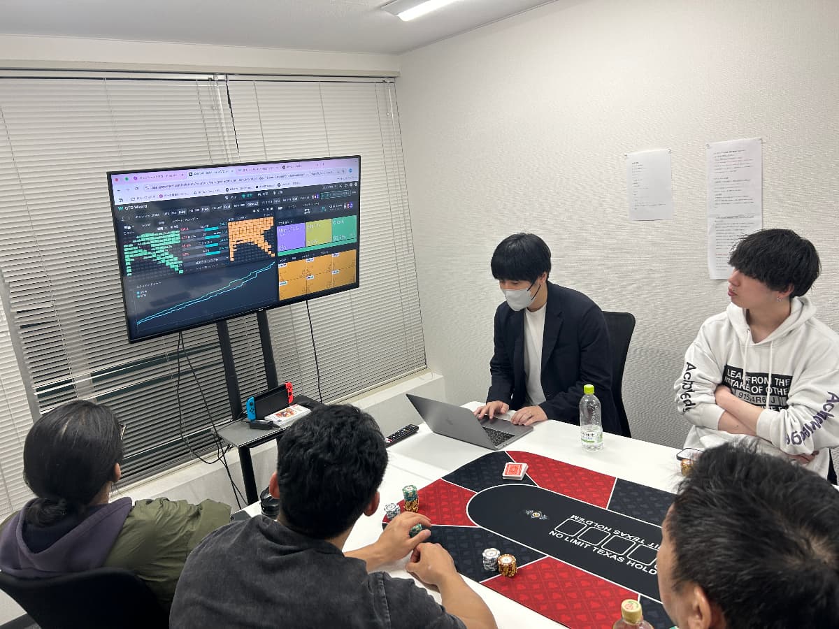 コミットポーカースクールの授業の様子。講師がモニターに戦略ツールを映し、受講生がポーカーマットを囲んで話を聞いている